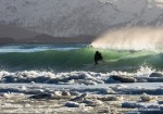 Alaskan surfer Gart Curtis rides a wave with snow, slush, and ice packed against the beach during a cold winter surf session in Homer, Alaska. The snowy Kenai mountain range rises from Kachemak Bay in the background.