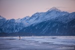 A surfer walking across a frozen beach after sunset with his surfboard.